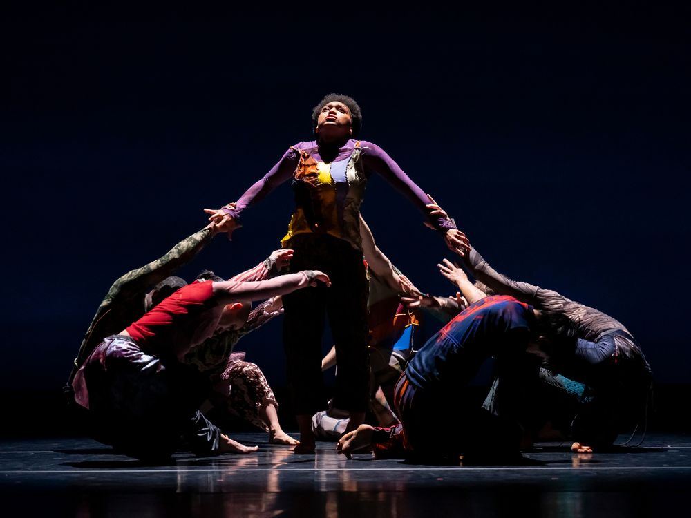 A group of dancers, in mis-matched clothes, are crouched on the ground, circling one Black dancer standing upright. The dancer's face is tilted up to the ceiling, yet their arms are being pulled down by the grabbing hands of the rest of the group.