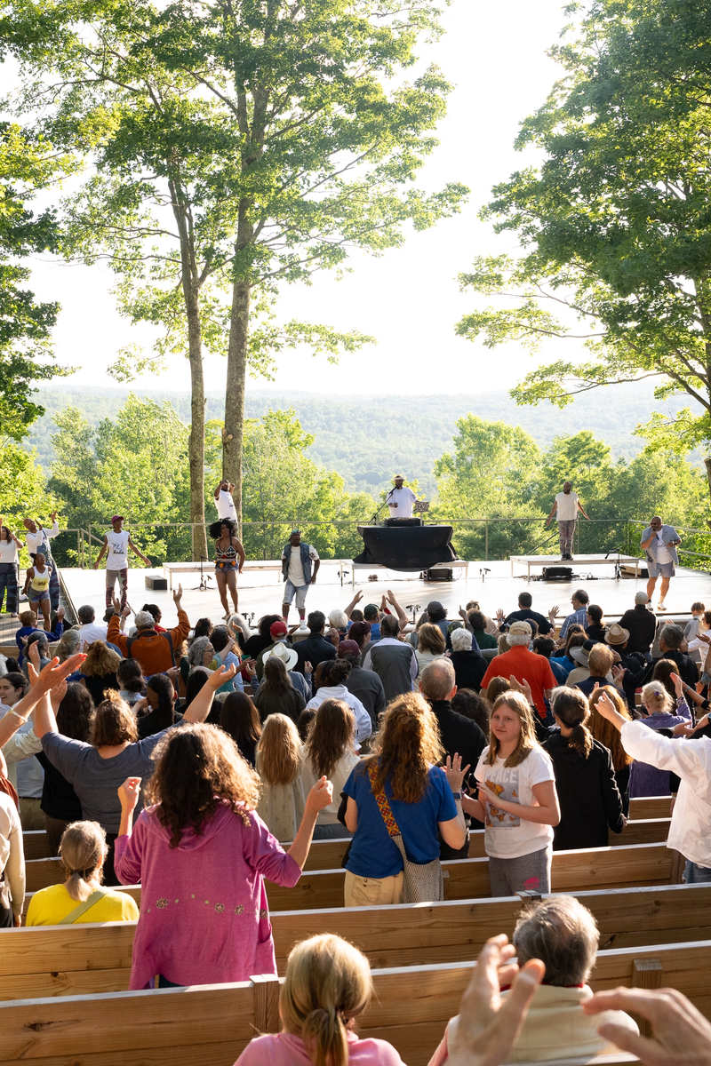 A full audience for the outdoor stage stands after watching a performance. The bright green scenery surrounds the audience and stage.